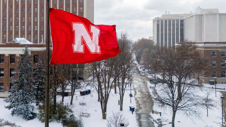 A red flag with a white Husker N waves over a sidewalk on the University of Nebraska–Lincoln's City Campus. Snow covers the ground.